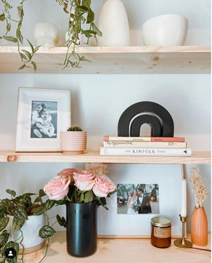 Two light wood shelves with white vases and a picture frame and a desk with salmon flowers on a black vase and a white vase with a plant on top.