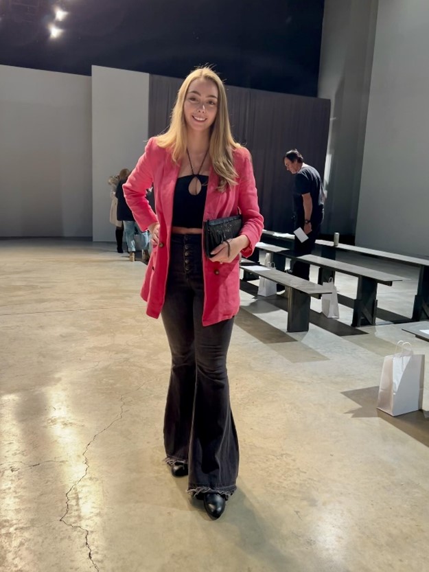 Women stading up wearing a pink blazer and black pants and top holding a small purse.