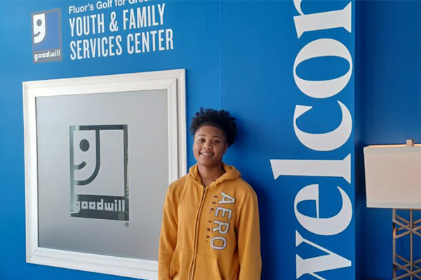 Women standing in front of a blue wall saying welcome and with the Goodwill logo. She is wearing a yellow sweatshirt and black pants.