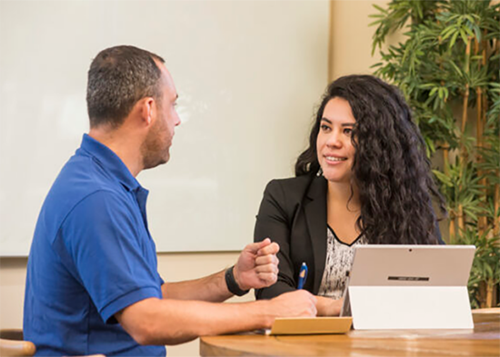 Men wearing a blue t-shirt is talking to a women with long curl brown hair wearing a black suit and white shirt. He is holding a pen and she has her computer in front of her.