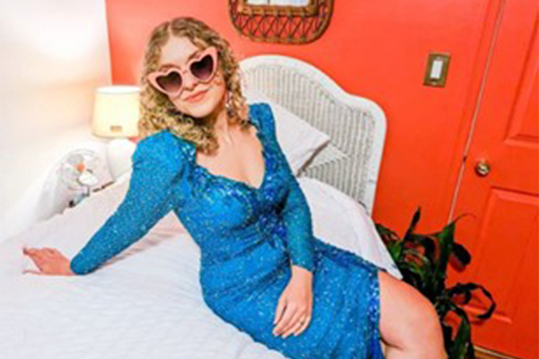 Women seating on a bed wearing a long blue sequin dress an sunglasses.