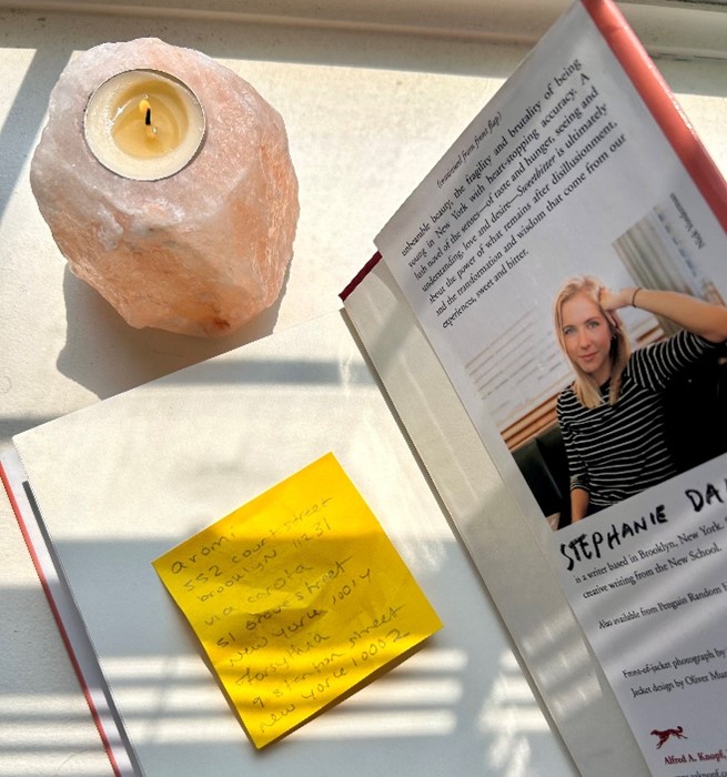An image of an open book with a yellow post stick in one side and the picture of the author on the other side with a lit candle on the side.