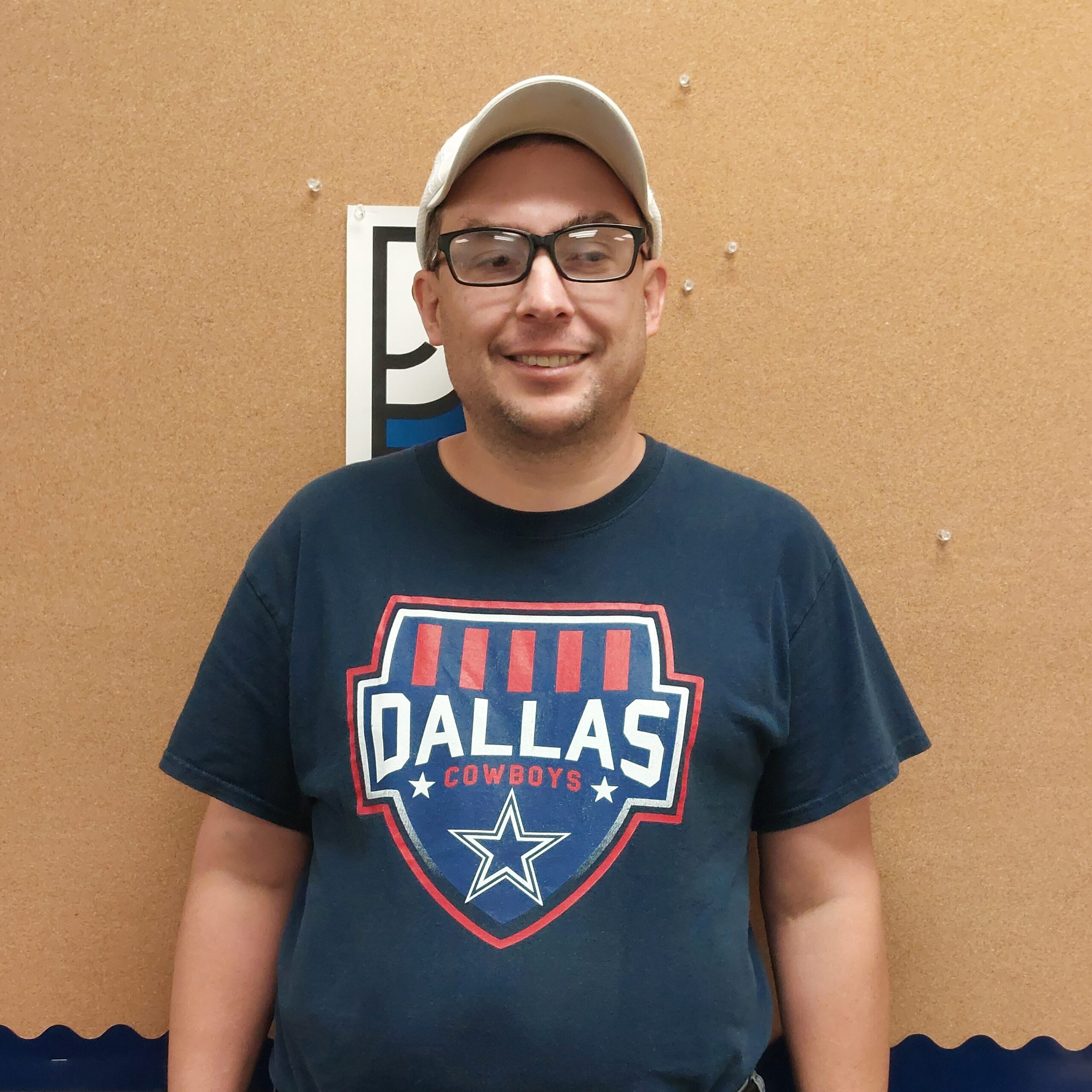 Image of a man standing in front of a orange wall. He wears reading classes, a cream hat and a Dallas navy blue shirt.