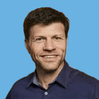 Portrait of a smiling man with short dark hair in a blue shirt against a solid blue background.