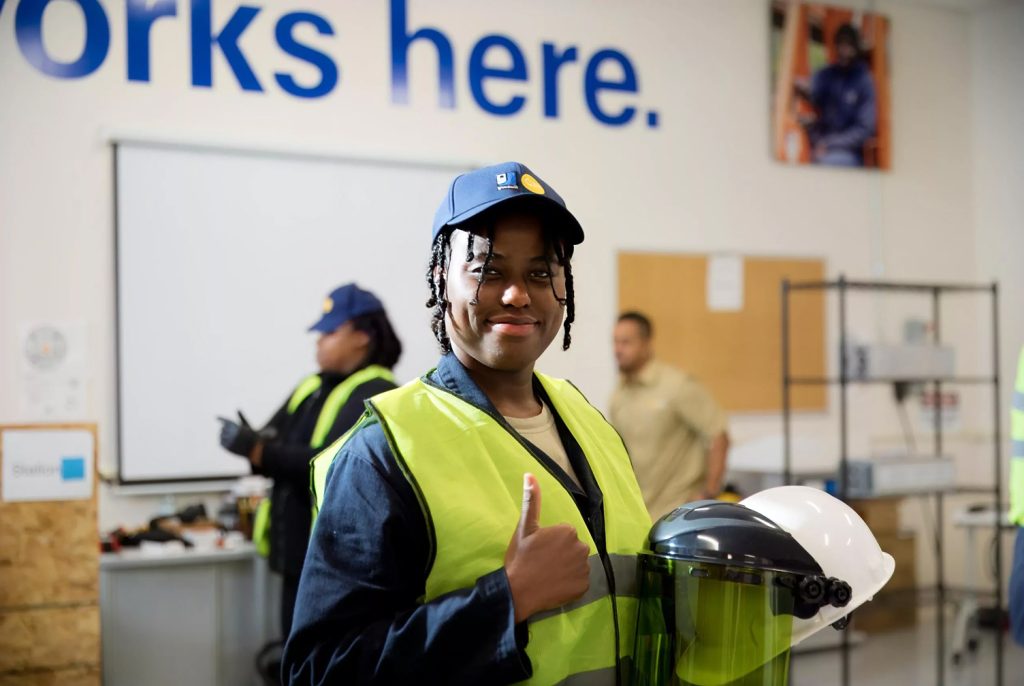 Smiling worker in a neon-yellow safety vest and blue cap gives a thumbs up inside a workshop.