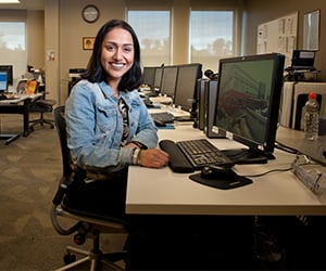 Smiling woman in a denim jacket sits at a desk with multiple computer monitors in a bright office.