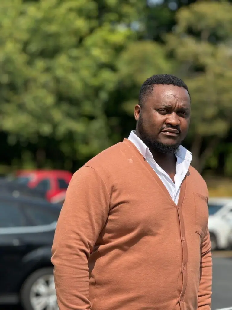 Portrait of a man wearing a peach cardigan in a parking lot with trees behind him.