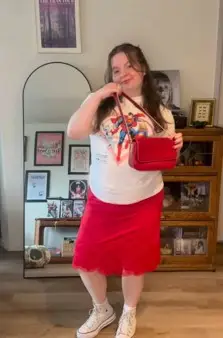 A women wearing a white T and red mid length skirt holding a red purse standing in a room with a tall mirror and a shelf in the back