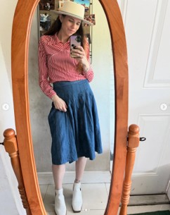 Women wearing a hat, red long sleev shirt, blue knee length skirt and white tennis shoes standing in front of a wooden round mirror